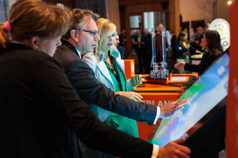 Een groep mensen staat bij een interactieve tentoonstellingsruimte. Een man en een vrouw kijken geconcentreerd naar een groot touchscreen, waar ze samen aan werken. Ze bevinden zich in een druk bezochte ruimte met oranje kleuraccenten en technologieën op de achtergrond. De sfeer lijkt geïnspireerd door innovatieve ontwikkelingen. De foto is genomen door Jan Buwalda in Groningen.