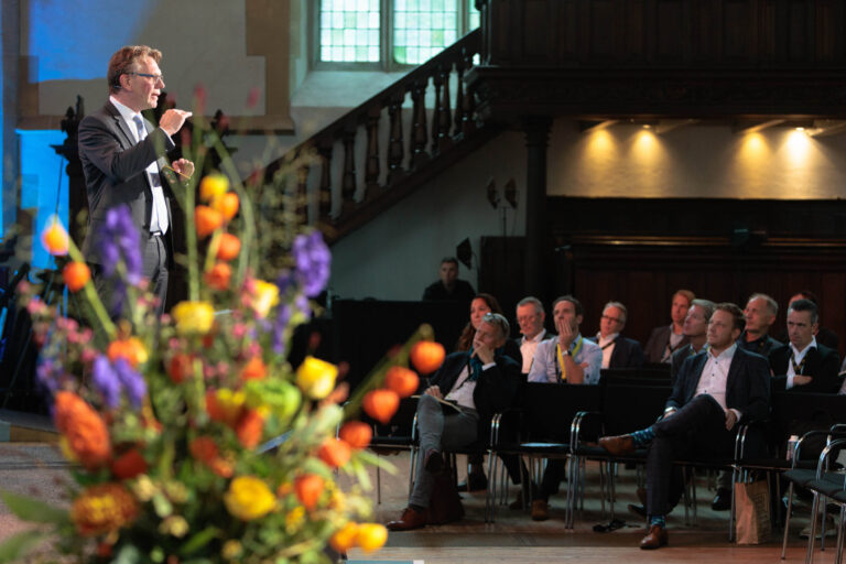 Een spreker staat voor een aandachtig publiek in een zaal met houten details en grote ramen. Op de voorgrond is een kleurrijk boeket bloemen te zien, wat een sfeervolle toevoeging vormt aan de setting. De aanwezigen lijken geboeid te luisteren naar de presentatie. Fotograaf: Jan Buwalda.