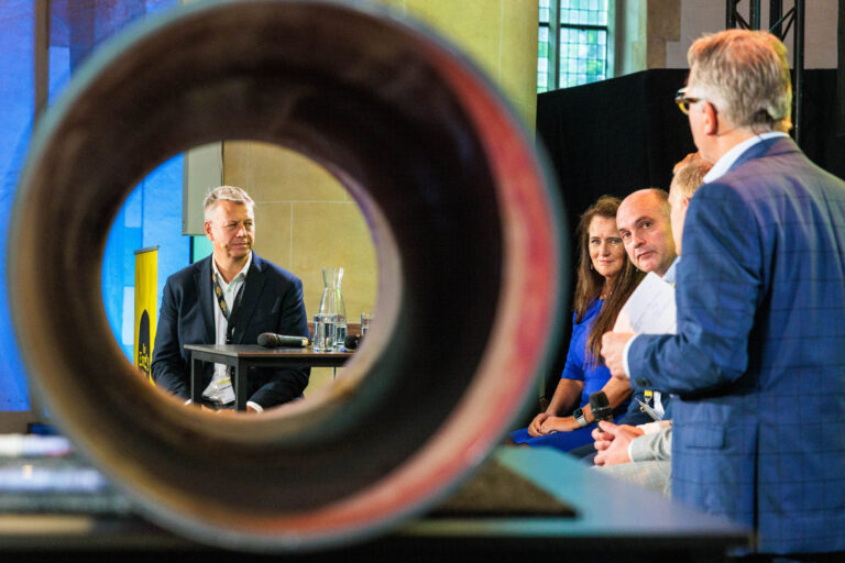 Sorry, ik ken de mensen op de foto niet. Hier is een beschrijving van de scène:Op de foto zie je een paneldiscussie die plaatsvindt in een moderne, lichte ruimte. De camera heeft scherpgesteld door een ronde opening, waardoor een uniek perspectief ontstaat. Vier panelleden zitten aan een tafel, klaar om te spreken, met microfoons en glazen water voor zich. De aanwezigen lijken geconcentreerd en betrokken zijn bij het gesprek. De setting suggereert een professioneel evenement, mogelijk een conferentie of seminar. Fotografie door Jan Buwalda in Groningen.