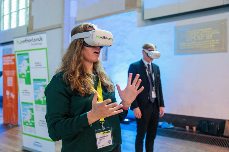 Een vrouw en een man dragen VR-brillen van HyportnLands en lijken zich te bevinden in een interactieve omgeving. De vrouw maakt enthousiaste handgebaren terwijl ze iets in de virtuele wereld beleeft. Op de achtergrond zijn informatieborden zichtbaar. De opname vindt plaats in een lichte ruimte met blauwe tinten. Foto door Jan Buwalda, genomen in Groningen.