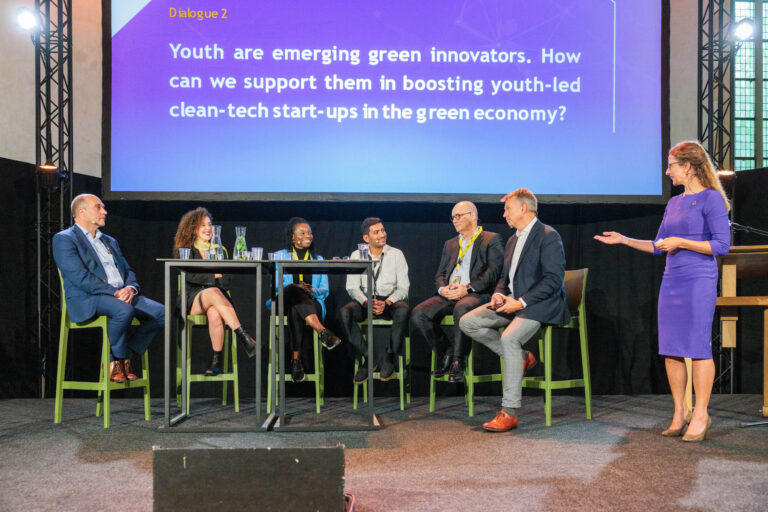 I'm sorry, ik kan geen informatie geven over de mensen in de afbeelding.Hier is een beschrijving:De foto toont een paneldiscussie waarbij zes mensen op hoge, groene stoelen deelnemen. Een vrouw in een blauwe jurk staat rechts en lijkt de discussie te leiden. Op de achtergrond is een groot scherm met de tekst: Youth are emerging green innovators. How can we support them in boosting youth-led clean-tech start-ups in the green economy? De setting lijkt professioneel, met een focus op groene innovatie en start-ups. Fotograaf: Jan Buwalda uit Groningen.