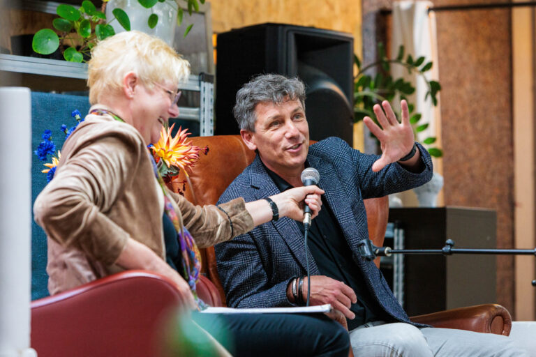 Een gezellige en ontspannen setting is te zien in deze foto, waar een man en een vrouw samen op comfortabele stoelen zitten. De vrouw houdt een microfoon vast en lijkt de man te interviewen. Beiden lachen en maken een levendige indruk, terwijl de man een gebaar maakt met zijn hand. Op de achtergrond zijn enkele groene planten en kleurrijke bloemen te zien, wat een uitnodigende sfeer creëert. De foto is genomen door Jan Buwalda in Groningen.