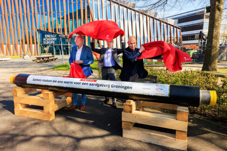 Dit is een foto genomen door Jan Buwalda in Groningen, waar drie mannen een ceremonieel moment houden bij de onthulling van een pijp. De pijp heeft de tekst: Samen maken we ons warm voor een aardgasvrij Groningen. Dit gebeurt voor het gebouw van de Energy Academy Europe. De mannen verwijderen rode doeken van de pijp, symbool van een nieuwe stap richting duurzame energie. Het gebouw op de achtergrond heeft een moderne en duurzame architectuur met veel glas en hout.