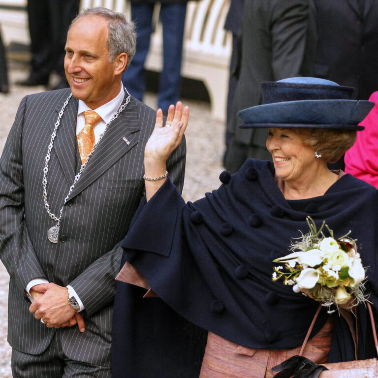 Het beeld toont een man in een gestreept pak met een ceremonieketting, naast een vrouw die een elegante zwarte hoed en bijpassende mantel draagt. Ze glimlacht en lijkt te wuiven naar een menigte, terwijl ze een boeket bloemen vasthoudt. De sfeer is feestelijk en plechtig, wat suggereert dat het gaat om een officiële bijeenkomst of ceremonie. De foto is genomen door Jan Buwalda in Groningen.