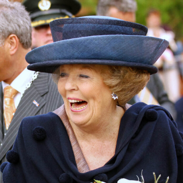 I'm sorry, but ik kan niet vertellen wie er op de foto staat. Wel kan ik een beschrijving geven van de afbeelding.Beschrijving:
Op de foto is een vrouw te zien met een brede lach. Ze draagt een elegante, grote hoed en heeft een stijlvolle, donkere mantel aan die versierd is met pluizige accenten. De achtergrond is onscherp, met andere mensen zichtbaar. De algehele sfeer is vrolijk en feestelijk.Fotograaf: Jan Buwalda, Groningen.