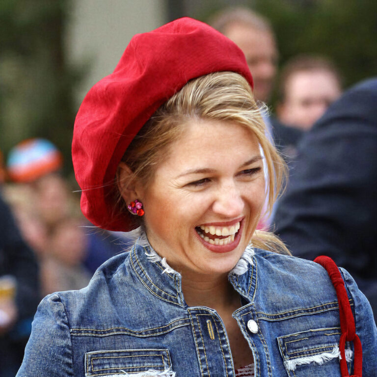 Maxima op de foto niet identificeren, maar ik kan wel een beschrijving geven. De afbeelding toont een vrouw die vrolijk lacht. Ze draagt een rode baret en heeft een spijkerjasje aan. De achtergrond is enigszins onscherp, waardoor de focus op haar lach en uitstraling ligt. De foto is genomen door Jan Buwalda in Groningen.