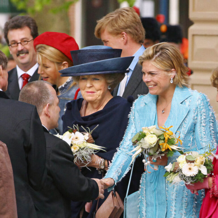 Het beeld toont een groep mensen in een feestelijke setting. Twee vrouwen staan op de voorgrond: een oudere vrouw met een elegante donkerblauwe hoed en een jongere vrouw in een lichtblauw ensemble. Ze dragen allebei boeketten bloemen en lachen naar de mensen voor hen. De sfeer is feestelijk en hoffelijk. Dit moment is vastgelegd door de fotograaf Jan Buwalda in Groningen.