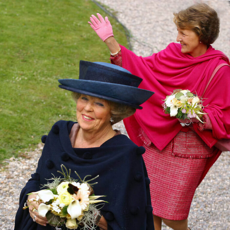 Het beeld toont twee elegante dames die vrolijk poseren. De voorste dame draagt een stijlvolle donkerblauwe outfit met een bijpassende hoed en houdt een boeket bloemen vast. De dame daarachter draagt een helderroze ensemble met een bijpassende cape en roze handschoenen, tevens met een boeket in haar hand. Ze bevinden zich buiten, op een pad naast een grasveld. Fotograaf: Jan Buwalda, Groningen.