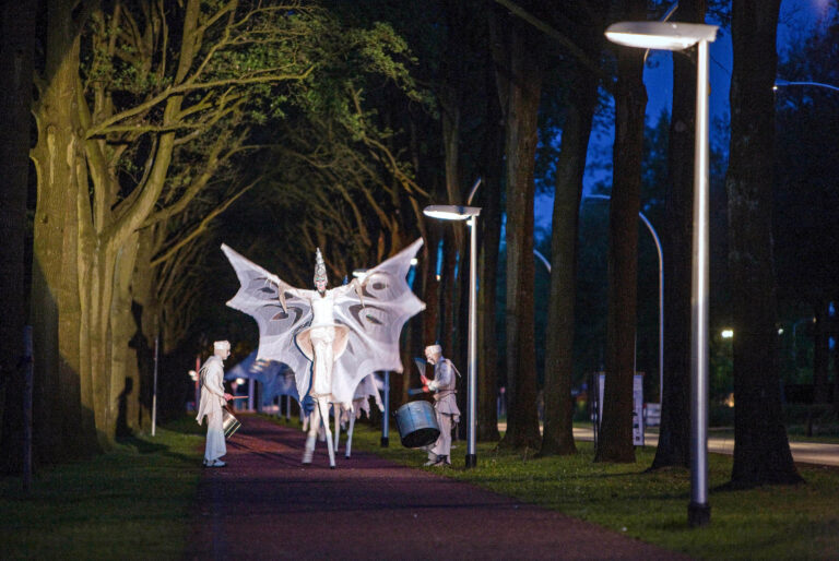 Dit betoverende nachtelijke tafereel toont een performer op stelten, gekleed als een majestueuze vlinderfiguur, die over een bomenlaan in Groningen loopt. Omringd door muzikaal ondersteunende figuren, baadt de scène in zacht licht, waardoor een magische sfeer ontstaat. De bomen vormen een tunnelachtige setting, die de aandacht naar het middelpunt van de act trekt. Deze foto is gemaakt door Jan Buwalda.