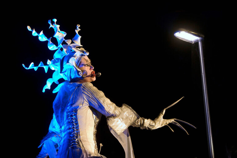 Een performer is dramatisch verlicht in een nachtelijke scène. De persoon draagt een opvallend kostuum met scherpe, gedraaide hoornachtige versieringen op het hoofd, glinsterend in blauw licht. Ze hebben een microfoon bij hun mond en steken een hand met lange, sierlijke vingers uit naar een felle lichtbron boven hen. Het contrast tussen het donkere omgevingslicht en de heldere belichting creëert een mystieke sfeer. Foto door Jan Buwalda, Groningen.