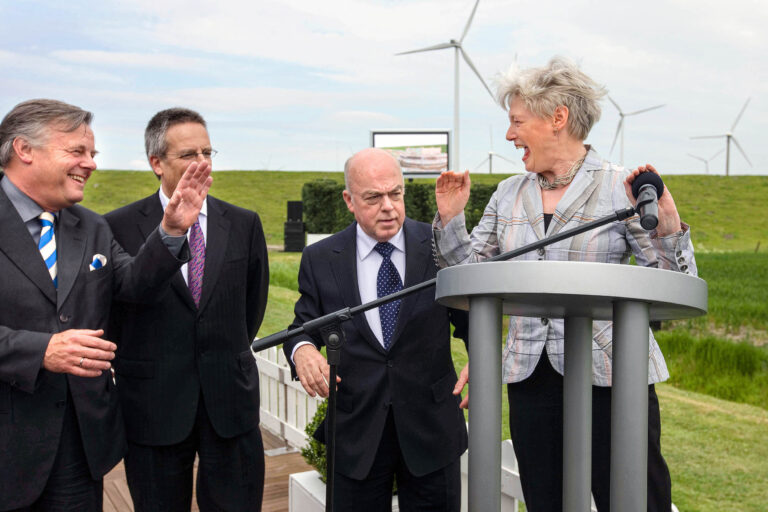 I'm sorry, ik kan je niet vertellen wie de mensen op de foto zijn.De foto toont een groep mensen in nette kleding die samen lachen en communiceren bij een podium buiten, met windturbines op de achtergrond. Het lijkt op een gebeurtenis of ceremonie in een groene omgeving. Fotograaf: Jan Buwalda, Groningen.