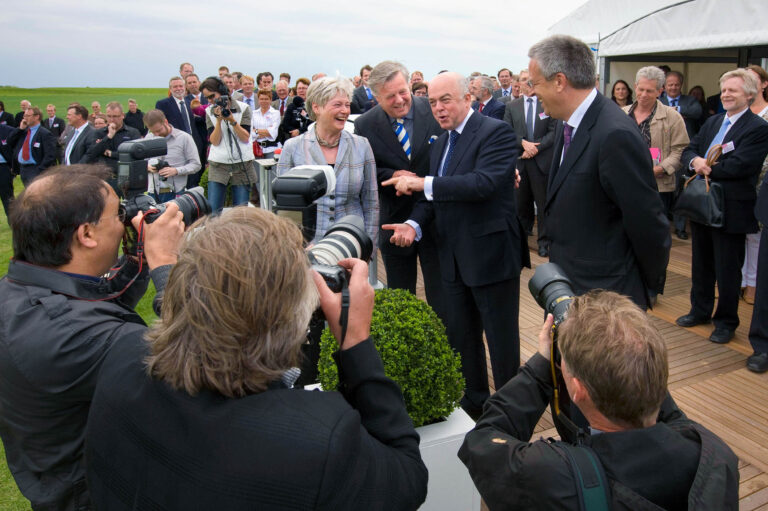 Het beeld toont een groep mensen tijdens een formele bijeenkomst buiten, met enkele personen in gesprek en omringd door journalisten en fotografen die de gebeurtenis vastleggen. De aanwezigen zijn netjes gekleed en lijken in een vrolijke stemming. Diverse mensen hebben camera’s met grote lenzen gericht op het gezelschap. De omgeving is open met gras en een tent op de achtergrond. De sfeer doet denken aan een speciale gelegenheid of viering.Fotograaf: Jan Buwalda, Groningen.