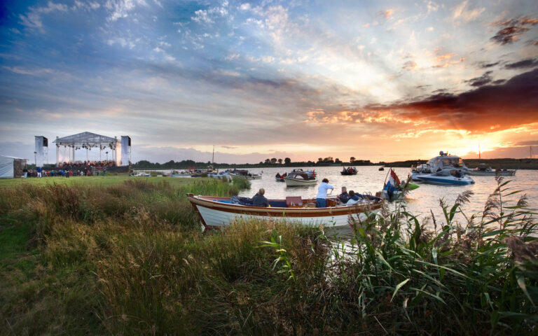 Dit sfeervolle beeld toont een prachtig buitenconcert aan het water bij zonsondergang, gefotografeerd in Groningen. Op de voorgrond zijn mensen te zien die ontspannen in een boot zitten, terwijl op de achtergrond een verlicht podium staat waar het concert plaatsvindt. De lucht is kleurrijk dankzij de ondergaande zon, wat een betoverende sfeer creëert. Fotograaf: Jan Buwalda.