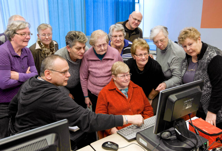 Dit beeld toont een groep senioren die aandachtig naar een computerscherm kijken terwijl een man hen iets uitlegt. De meeste mensen staan rechtop om het scherm beter te kunnen zien, en er is een vrouw die zit en naar het scherm kijkt. De sfeer is nieuwsgierig en leergierig. Deze foto is gemaakt door Jan Buwalda in Groningen.