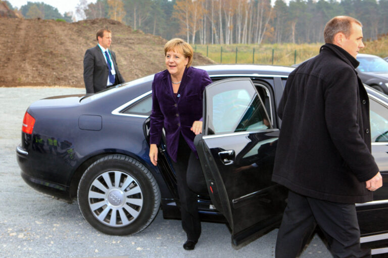 Een persoon stapt uit een zwarte Audi op een buitenlocatie. De auto staat geparkeerd op grind en op de achtergrond zijn enkele bomen te zien. Een man in een donkere jas staat naast de auto. In de verte staat een andere man in pak met een badge om zijn nek. De foto is genomen door Jan Buwalda in Groningen.