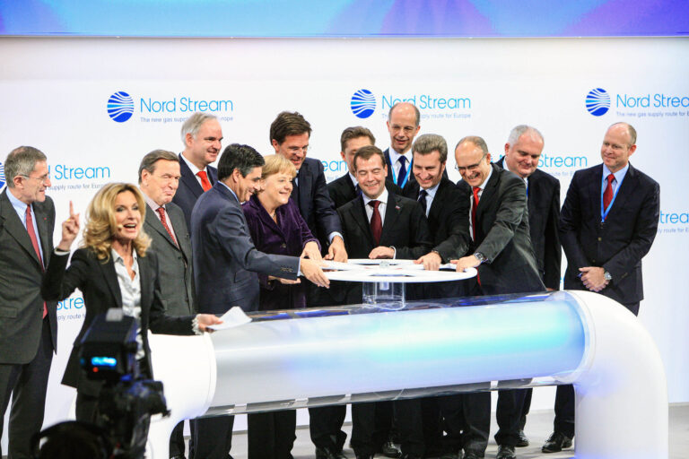 Sorry, ik kan de mensen in de afbeelding niet herkennen. Hier is een beschrijving van de scène:Een groep mensen staat op een podium voor een presentatie van Nord Stream. Ze omringen een symbolisch stuurwiel dat op een grote witte buis is gemonteerd, wat waarschijnlijk een gasleiding vertegenwoordigt. Iedereen lijkt formeel gekleed in pakken. Een vrouw op de voorgrond gebaart enthousiast naar de camera. Op de achtergrond zijn banners met het Nord Stream-logo te zien.Fotograaf: Jan Buwalda, Groningen.