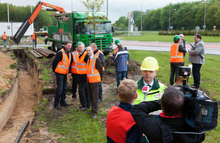 De afbeelding toont een groep mensen op een bouwplaats in Groningen. Er zijn mannen in oranje veiligheidshesjes die naast een uitgegraven sleuf staan. Op de achtergrond is een groene vrachtwagen met een grijpkraan zichtbaar. Voorin is er een man in een geel veiligheidsharnas en helm die geïnterviewd wordt door een journalist met een microfoon van RTV Drenthe. Rondom deze scène zijn andere mensen druk in gesprek of bezig met werkzaamheden. De omgeving oogt als een bouwproject langs de weg, met groene bomen in de verte.Fotograaf: Jan Buwalda.