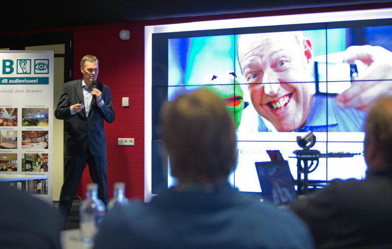 Een spreker in een blauwe blazer presenteert met een microfoon in de hand tijdens een bijeenkomst. Op de achtergrond is een groot scherm te zien met een opvallende afbeelding van een lachende man en Kermit de Kikker. Links van het scherm staat een banner van dB audiovisueel met foto's van verschillende presentatiesystemen. De setting lijkt een professionele presentatieomgeving te zijn met toehoorders op de voorgrond. Fotograaf: Jan Buwalda, Groningen.