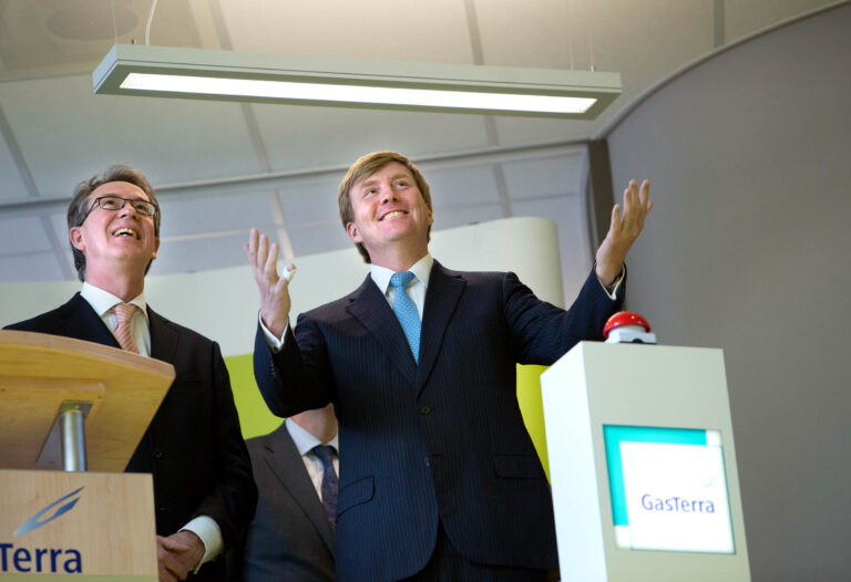 Ik kan de personen op de foto niet identificeren, maar ik kan wel een beschrijving geven.Op de foto staan twee mannen in nette pakken in een zakelijke omgeving. Ze kijken beiden glimlachend naar boven. Voor de man rechts staat een podium met een rode knop en een logo van GasTerra. Het plafond en een hanglamp zijn op de achtergrond zichtbaar. Deze foto is genomen door Jan Buwalda in Groningen.