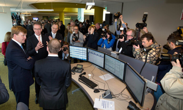 In deze foto, genomen door Jan Buwalda in Groningen, zien we een groep mensen die zich verzameld heeft rond een persoon die spreekt. Op de voorgrond staan meerdere computerschermen op een bureau. Verschillende fotografen drukken enthousiast op hun camera's om het moment vast te leggen, terwijl andere aanwezigen aandachtig luisteren en toekijken. De ruimte is modern ingericht, met een opvallend geel element op de achtergrond, en er heerst een bedrijvige, professionele sfeer.