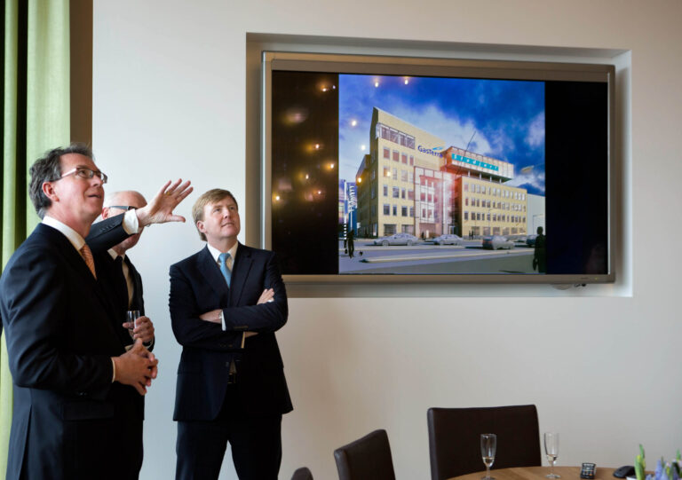 Dit is een foto van drie mannen in pakken die aandachtig naar een scherm kijken waarop een bouwproject van GasTerra te zien is. De setting is een moderne vergaderruimte met een grote presentatie op het scherm. Een van de mannen wijst naar de presentatie. De glazen op tafel suggereren dat het een formele bijeenkomst of presentatie is. Fotograaf: Jan Buwalda, Groningen.