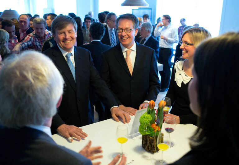 Het beeld toont een groep mensen die bijeen zijn voor een evenement, gezellig pratend rondom een tafel. Ze dragen formele kleding, wat wijst op een zakelijke of speciale gelegenheid. Op de tafel staan glazen met drankjes en een bloemstuk als decoratie. De sfeer lijkt ontspannen en joviaal. Fotograaf: Jan Buwalda uit Groningen.