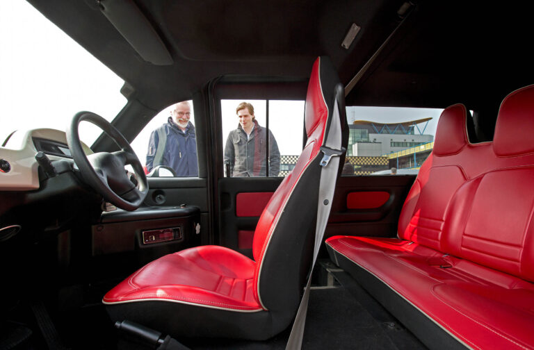 Dit beeld toont het interieur van een voertuig met opvallende rode lederen bekleding op de voor- en achterbank. De foto biedt een blik van binnenuit, waarbij twee personen nieuwsgierig van buitenaf naar het voertuig kijken. Het moderne ontwerp van de autostoelen wordt mooi uitgelicht, en op de achtergrond is een gebouw zichtbaar dat doet denken aan stadse architectuur. Deze foto is genomen door Jan Buwalda in Groningen.