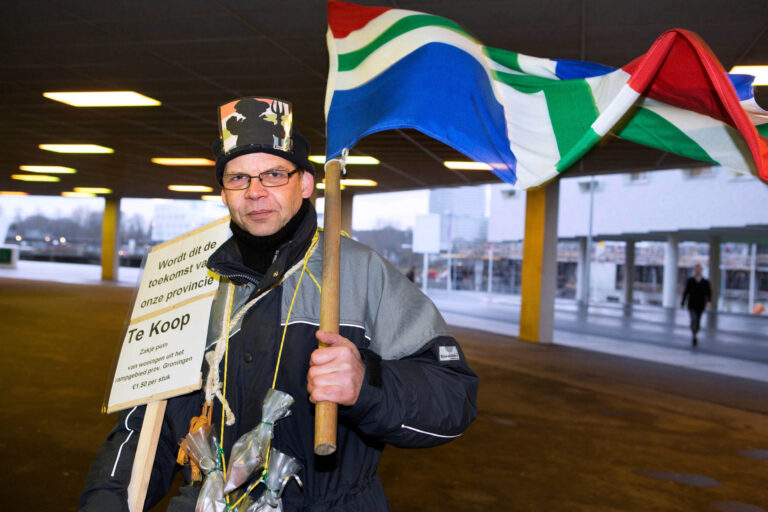 Op de foto is een man te zien die een kleurrijke vlag vasthoudt en een bord draagt met de tekst: Wordt dit de toekomst van onze provincie? Te Koop. Zakje puin van woningen uit het rampgebied prov. Groningen €1,50 per stuk. De man draagt een hoed met een tekening en heeft kleine zakjes puin bij zich. De achtergrond toont een stadssfeer met moderne architectuur. De opname is gemaakt door fotograaf Jan Buwalda in Groningen.