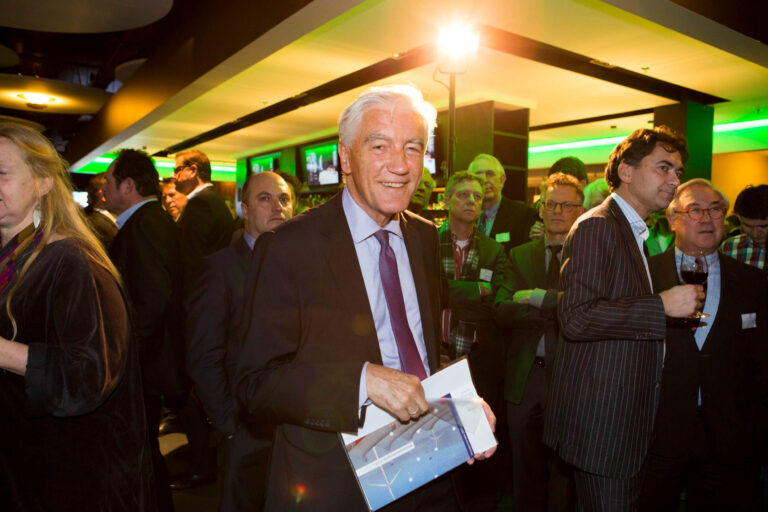 Ik kan helaas geen mensen in de foto herkennen.Beschrijving: De foto toont een drukke bijeenkomst in een moderne ruimte met levendige groene verlichting. Een oudere man staat middenin, gekleed in een donker pak met een lichtblauwe stropdas, glimlachend naar de camera terwijl hij een folder vasthoudt. Om hem heen staan meerdere mannen, gekleed in formelere outfits, en een vrouw met lang haar in een donker kledingstuk. De sfeer lijkt sociaal en professioneel.Fotograaf: Jan Buwalda, Groningen.