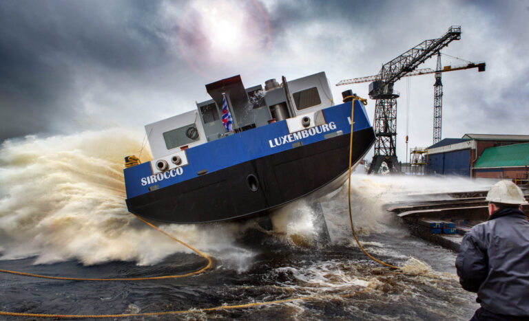 Dit dynamische beeld toont de spectaculaire tewaterlating van een schip, genaamd Sirocco Luxembourg, in de haven van Groningen. De kracht van het water spettert hoog op terwijl het schip het water raakt, en op de achtergrond zijn havenkranen zichtbaar onder een bewolkte, dramatische lucht. Een man in de voorgrond kijkt toe terwijl het schip zijn weg vindt naar het water. Fotograaf: Jan Buwalda.