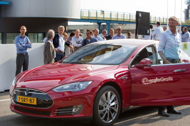 Een groep mensen staat buiten bij een moderne faciliteit nabij een rode Tesla, met het Energielabel op de auto. Een man lacht terwijl hij de portier van de auto opent. De groep lijkt in gesprek. Deze foto is genomen door Jan Buwalda in Groningen.
