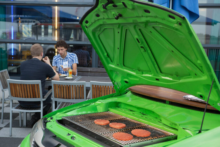 Een creatieve en unieke foto gemaakt door Jan Buwalda in Groningen toont een groene auto met de motorkap open, waarin een barbecue is ingebouwd. Op de grill liggen verschillende hamburgers te bakken. Op de achtergrond zitten twee mensen aan een tafel buiten, waar ze genieten van een drankje en elkaars gezelschap. Deze innovatieve setting combineert auto-entertainment met culinaire ervaringen, waardoor een speelse en gezellige sfeer wordt gecreëerd.