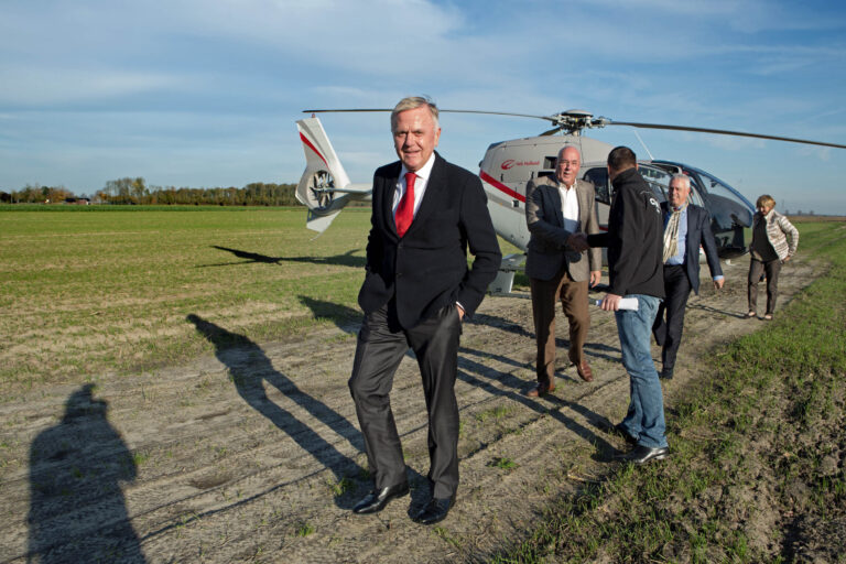 Op de foto gemaakt door Jan Buwalda in Groningen is een groep mensen te zien die net uit een helikopter zijn gestapt. Ze staan op een landelijk grasveld onder een heldere blauwe hemel. Eén van de mannen, gekleed in een donker pak met een rode stropdas, loopt naar voren terwijl anderen op de achtergrond met elkaar praten of elkaar de hand schudden. De schaduwen van de mensen en de helikopter zijn duidelijk zichtbaar op de grond.