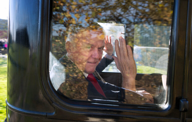 In deze foto, genomen door Jan Buwalda in Groningen, zien we een ouder persoon die vanuit een voertuig naar buiten zwaait. De reflecties in het raam tonen een serene omgeving met herfstbladeren, wat een warme sfeer toevoegt aan de opname. De persoon lijkt ontspannen en vriendelijk terwijl hij naar de camera kijkt. De lichtinval en schaduwen geven de foto een levendig en uitnodigend karakter.