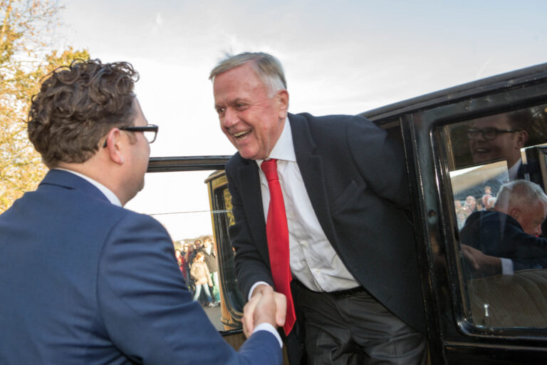 Een man in een zwart pak met een rode stropdas stapt glimlachend uit een auto en schudt de hand van een andere man in een blauw pak en bril. Op de achtergrond is een menigte mensen te zien die toekijkt. De foto straalt een gevoel van warmte en vrolijkheid uit. Fotograaf: Jan Buwalda, Groningen.