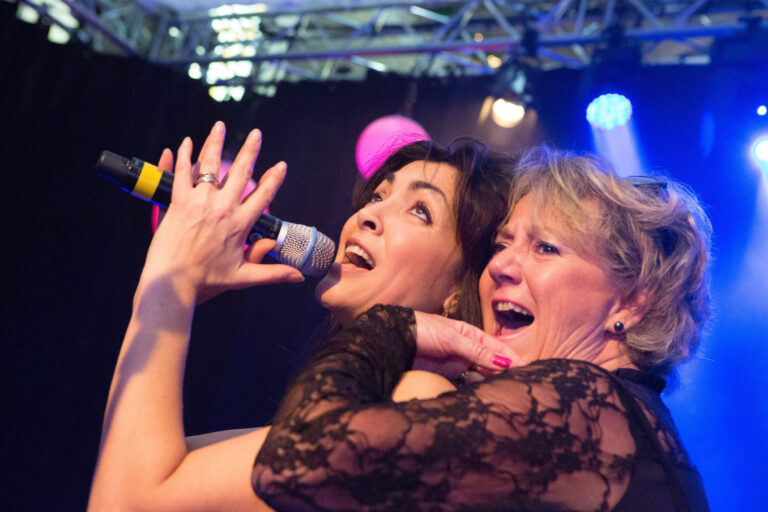 Een levendige afbeelding van twee vrouwen die samen zingen op een podium. De vrouw links houdt een microfoon vast en zingt vol overgave, terwijl de vrouw rechts enthousiast meedoet. De kleurrijke verlichting en het enthousiasme van de vrouwen creëren een feestelijke sfeer. Fotograaf: Jan Buwalda, Groningen.