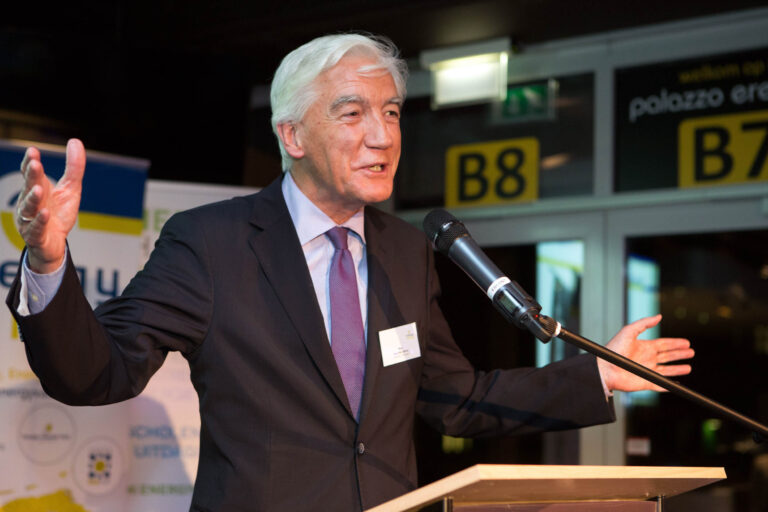 Het beeld toont een man in een donker pak en stropdas die een toespraak geeft achter een lessenaar. Hij spreekt gepassioneerd in een microfoon en maakt gebruik van handgebaren. Op de achtergrond zijn borden met opschriften zichtbaar. De foto is genomen door Jan Buwalda in Groningen.