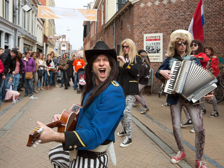 Op de foto, genomen door Jan Buwalda in Groningen, zien we een levendig straatoptreden. Een muzikale groep, gekleed in kleurrijke en flamboyante outfits, speelt energieke muziek terwijl ze door een drukke straat paraderen. Op de voorgrond staat een gitarist in een opvallend zwart-wit gestreepte broek en blauw jasje, die enthousiast zingt. Omstanders staan langs de straat, glimlachend en genietend van de performance, terwijl kinderen nieuwsgierig toekijken. Deze dynamische scène vangt de vrolijke en feestelijke sfeer van een stadsfestival.