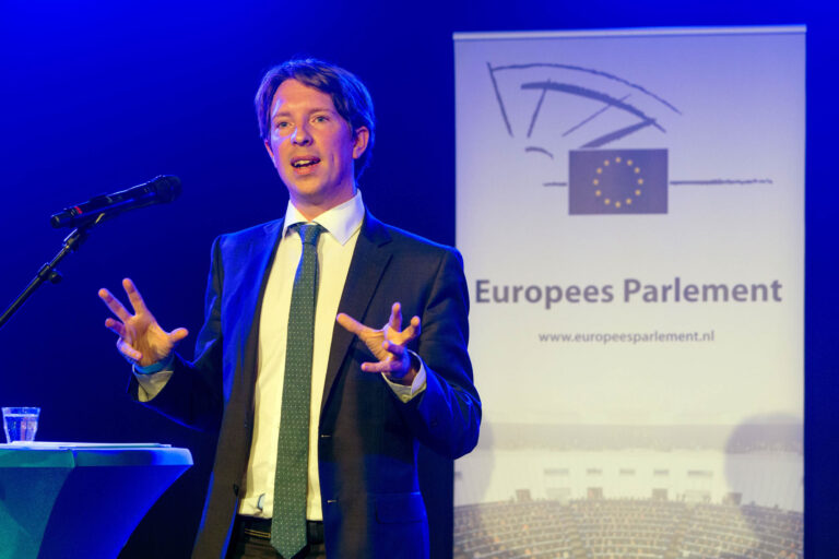 Het beeld toont een man in een pak die een presentatie geeft op een podium, naast een microfoon. Op de achtergrond staat een banner van het Europees Parlement. De verlichting rondom hem is blauw, wat een professionele sfeer creëert. Er staat een glas op tafel naast hem. Foto door Jan Buwalda, Groningen.
