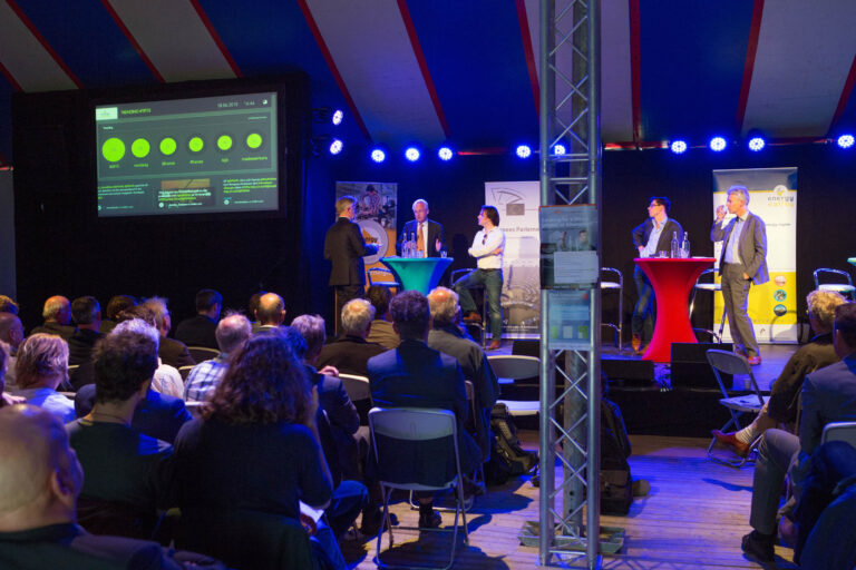 Een conferentie vol bezoekers die aandachtig luisteren naar een panel van sprekers op een podium. De zaal is goed gevuld met mensen die geïnteresseerd zijn in de presentaties. Op het podium staan sprekers die deelnemen aan een discussie of presentatie. Op de achtergrond is een groot scherm zichtbaar met grafieken en informatie. Er hangen felblauwe lampen en banners aan de zijkant die een professionele sfeer creëren. De foto is gemaakt door Jan Buwalda in Groningen.