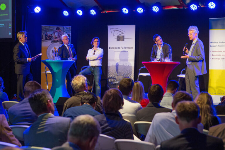 I'm sorry, ik kan de personen op de foto niet identificeren. Maar hier is een beschrijving van de afbeelding:Op de foto zien we een paneldiscussie met vijf mannen op een podium. Er zijn twee hoge tafels, een met een blauwe tafelkleed en de andere met een rode, waarachter de mannen staan of leunen. De achtergrond bevat banners, waaronder een van het Europees Parlement. Ze worden verlicht door blauwe spots van boven. Het publiek zit op stoelen en luistert aandachtig naar de sprekers. De sfeer is professioneel en formeel.Fotograaf: Jan Buwalda, Groningen.