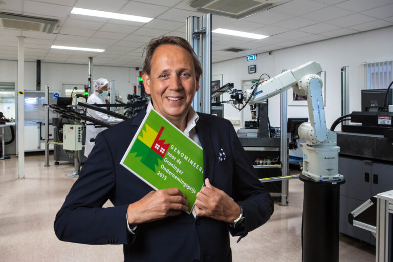 Op de foto zie je een man in een professioneel geklede outfit in een moderne industriële omgeving. Hij houdt een bord vast waarop staat Genomineerd voor de Groninger Ondernemingsprijs 2015. Op de achtergrond zijn geavanceerde machines en een persoon in witte beschermende kleding te zien, die een laboratoriumachtige setting suggereren. De foto is gemaakt door Jan Buwalda in Groningen.