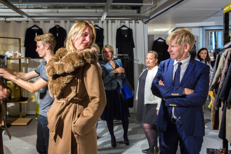 In een winkel staat een vrouw in een stijlvolle bruine jas met bontkraag in het middelpunt van de aandacht. Om haar heen staan mensen die geïnteresseerd toekijken. De ruimte is gevuld met kledingrekken en er heerst een levendige sfeer. De compositie vangt een moment van mode en sociale interactie. Foto door Jan Buwalda, Groningen.