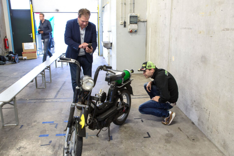 Op de foto is een man te zien die gebogen staat over zijn telefoon, terwijl een andere persoon een brommer van dichtbij bekijkt. Ze bevinden zich in een industriële ruimte met betonnen vloeren en muren. Een lange rij banken staat langs de zijkant, en er is een open deur zichtbaar waar een derde persoon langsloopt. De sfeer lijkt praktisch en ongedwongen. Fotografie door Jan Buwalda in Groningen.