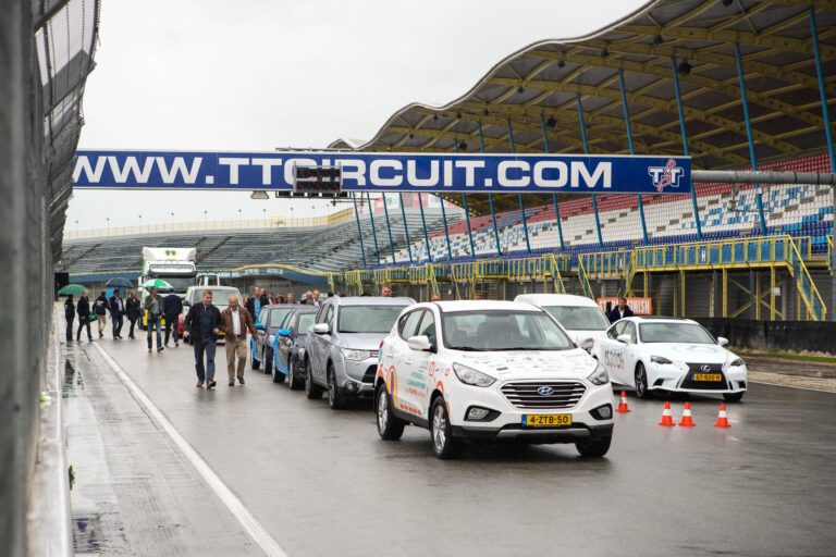 Dit beeld toont een regenachtige dag op het TT Circuit, waar verschillende auto's in een rij opgesteld staan op de pitstraat. Op de achtergrond zijn mensen te zien die over het natte asfalt lopen. De tribunes zijn leeg, en de overkoepelende structuur met een groot bord, WWW.TTCIRCUIT.COM, is goed zichtbaar. De sfeer is die van een voorbereidingsdag of een evenement dat op het punt staat te beginnen. Deze foto is gemaakt door fotograaf Jan Buwalda in Groningen.