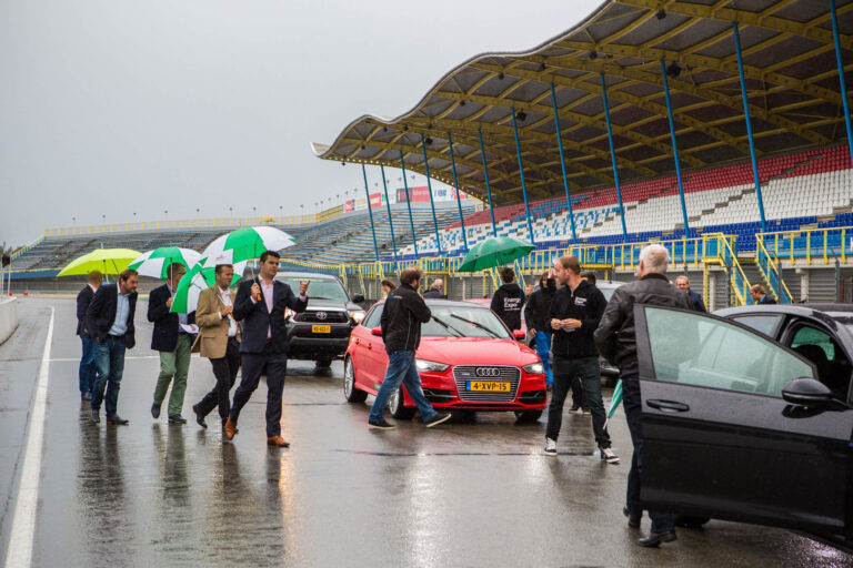 Op de foto is een groep mensen te zien die op een regenachtige dag op een racecircuit staan. Sommigen dragen paraplu's terwijl ze naast geparkeerde auto's staan, waaronder een rode Audi. De tribunes van het circuit zijn op de achtergrond zichtbaar. De mannen lijken in gesprek te zijn en er heerst een zakelijke sfeer. Deze foto is gemaakt door Jan Buwalda in Groningen.