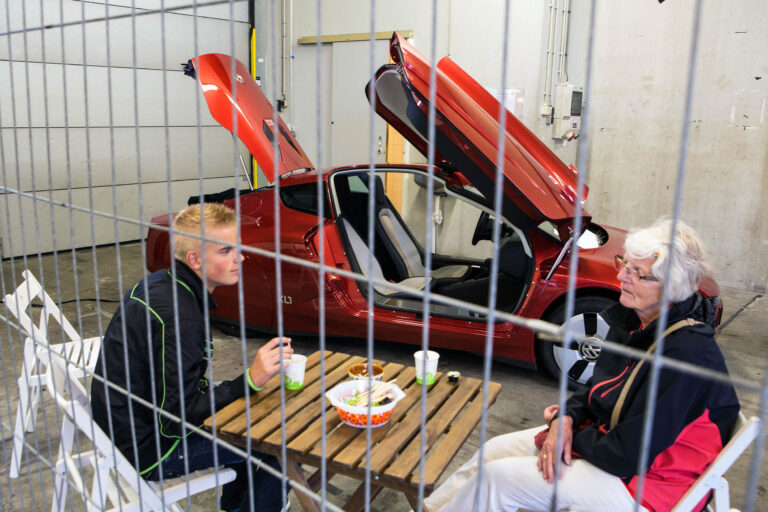 Een jonge man en een oudere vrouw zitten aan een houten tafel in een industrieel ogende ruimte. Op de tafel staan bekers en een schaal met lekkernijen. De tafel is opgesteld naast een opvallende rode auto met vleugeldeuren die openstaan. De setting wordt omheind door metalen tralies. De sfeer is informeel en ontspannen. Foto door Jan Buwalda, Groningen.