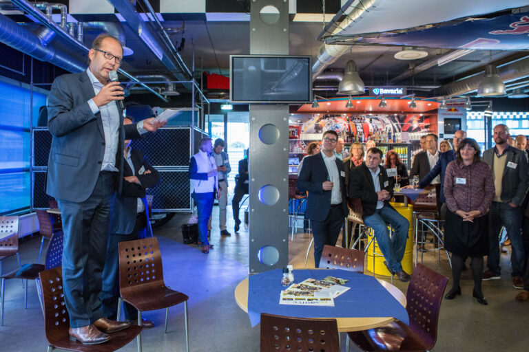 In een moderne, industriële setting met opvallende blauwe verlichting, zien we een man in pak die een toespraak houdt terwijl hij op een stoel staat. Hij spreekt voor een groep aandachtige mensen die om hem heen staan in een café-achtige omgeving. Er is een scherm en verschillende tafels en stoelen te zien. Het interieur heeft ook speelse en kleurrijke decoraties. De foto is genomen door Jan Buwalda in Groningen.