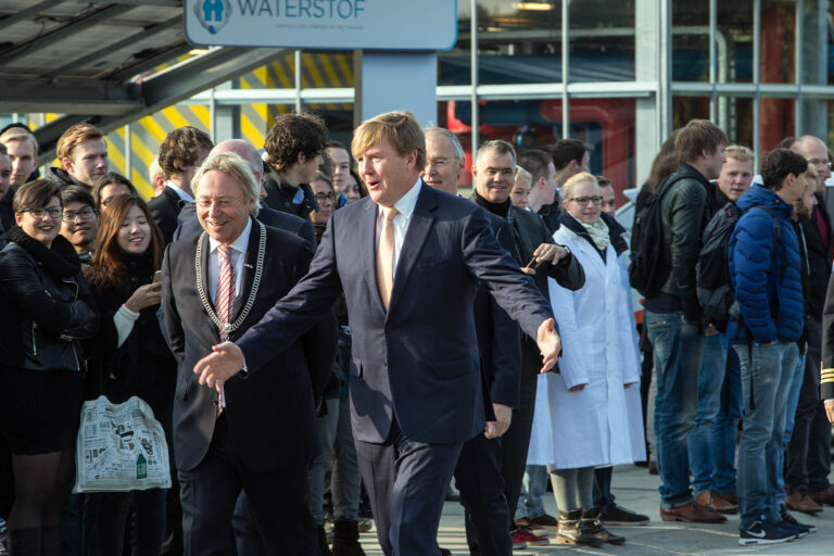 Deze foto, gemaakt door Jan Buwalda in Groningen, toont een groep mensen waaronder een man in een pak met een lichtgekleurde stropdas en een ambtsketen. Naast hem loopt een andere man in een donker pak met een oranje stropdas. Op de achtergrond staan toeschouwers, waaronder een persoon in een witte labjas, die het tafereel met belangstelling volgen. Boven hen is een bord met het woord Waterstof zichtbaar. De sfeer is levendig en formeel.