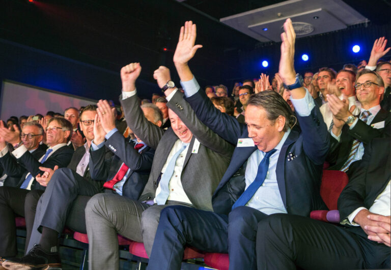 Dit beeld toont een groep goedgeklede mannen in een zaal, zittend in rode stoelen. Ze zijn enthousiast en klappen met hun handen in de lucht, wat duidt op een vreugdevol of inspirerend moment tijdens een evenement. De sfeer in de zaal is levendig, met theaterverlichting op de achtergrond en een menigte die de gebeurtenis bijwoont.Fotograaf: Jan Buwalda, Groningen.